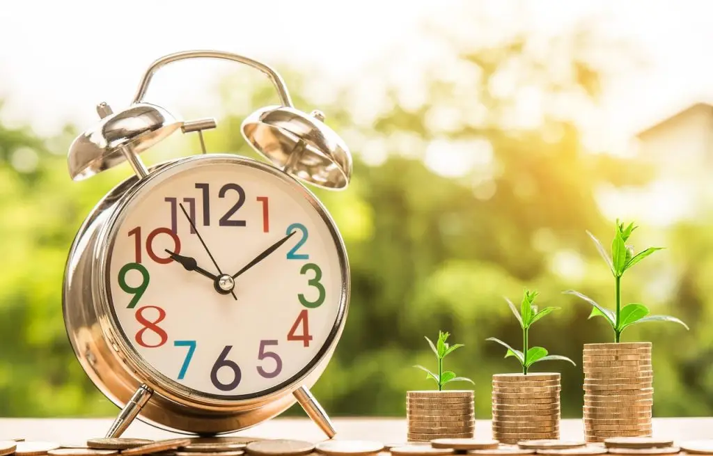 image of an old style alarm clock with multi coloured numbers next to coins and green shoots from small plants